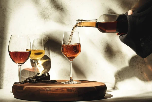 Wine tasting. Rose wine pouring into glass on background with selection of red, white and rose wines in glasses. Hard sunlight and shadows from foliage