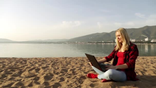 ビーチにノートパソコンを持つ女性。海と仕事だ。フリーランス。ソーシャルネットワーク夕方の海。砂浜だ。移動通信だ。遠隔作業だ。遠くの仕事だ。オンラインでチャット。楽しい仕事だ。ノートパソコン. — ストック動画