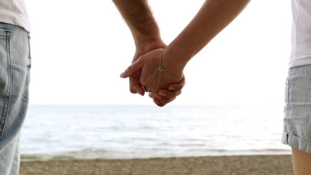 Par hålla händerna på bakgrund av havet. Närbild av händerna mot havet. Man och kvinna med händerna på stranden. Kärlek, romantik, nygifta. — Stockvideo