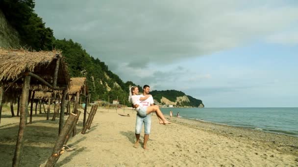 A man carrying a woman in his arms on the beach. Newlyweds at the sea.  Honeymoon, romance, just married. The groom carries his bride in his arms on the background of the sea. — Stock Video