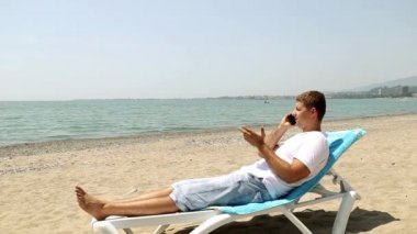 Telefon sahilde olan adam. Adam telefonda konuşurken bir şezlong üzerinde. Deniz arka planda telefonda konuşuyor. Güneşli bir plaj telefonda konuşan adam. 