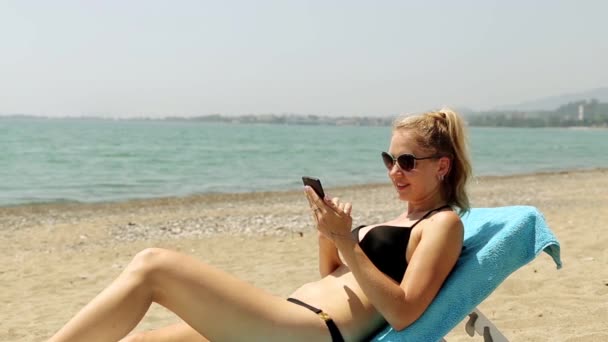 Kobieta na tle morza ze smartfonem w ręku, siedząc na Internecie. Dziewczyny na plaży w strój kąpielowy w Internecie z smartphone. Kobieta leżąc na leżaku z smatfonom. — Wideo stockowe
