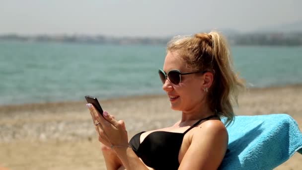 在泳装在海滩上与您的手机互联网的女孩。躺在躺椅上，智能手机的女人。随着智能手机在手，坐在互联网上的海面背景的女人. — 图库视频影像