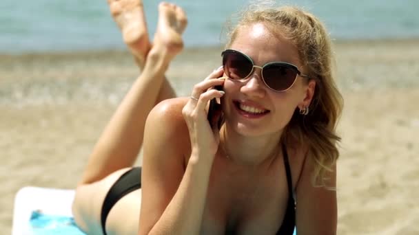 女性、サンラウン ジャーに横になっていると、電話で話しています。女性は電話で呼び出して、ビーチで日光浴、海でリラックスします。海の背景に電話で話している水着の女の子. — ストック動画