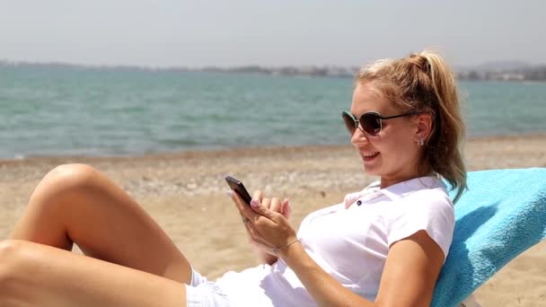 Kobieta w Internecie z smartphone na plaży. Dziewczyna z Internetu na tle morza. Kobieta odpoczynek nad morzem, patrząc na telefon. Kobieta leży na leżak ze smartfonem. — Wideo stockowe