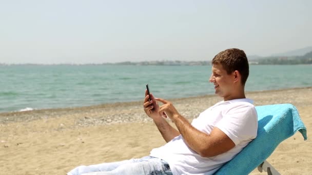 Hombre en Internet con smartphone en la playa. Hombre navegar por Internet en un fondo del mar. El hombre se encuentra en una tumbona con un teléfono inteligente. Hombre descansando en el mar, mirando el teléfono . — Vídeos de Stock