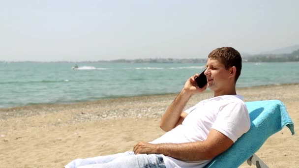 Hombre con teléfono en la playa. Un hombre en una tumbona hablando por teléfono. El hombre habla por teléfono en el fondo del mar . — Vídeos de Stock