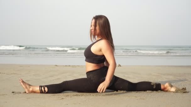Приваблива гнучка жінка сидить на шпагаті на морському пляжі — стокове відео