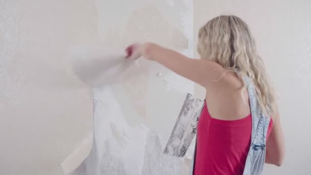 Uma mulher arranca o velho papel de parede da parede e faz reparos na casa — Vídeo de Stock