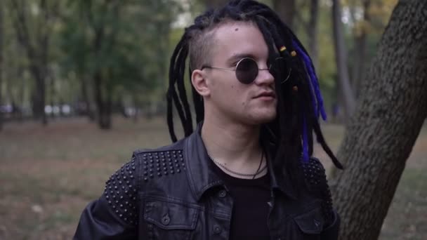 Retrato de un hombre con estilo en una chaqueta de cuero negro, gafas redondas y cabello con rastas — Vídeos de Stock