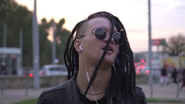 Portrait of a stylish man in a leather jacket, round glasses and dreadlocks. Fashionable teenager walks and listens to music with headphones — Stock Video