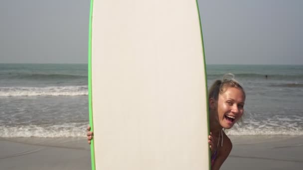 Femme drôle gaie se cache et regarde par derrière une planche de surf — Video
