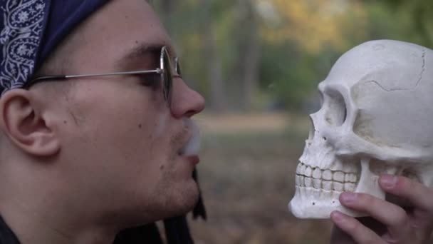 Человек выдыхает сигаретный дым в лицо черепа — стоковое видео