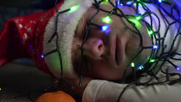Un hombre con un sombrero de Santa Claus y una guirnalda en su cara duerme en el suelo bajo el árbol de Navidad — Vídeos de Stock
