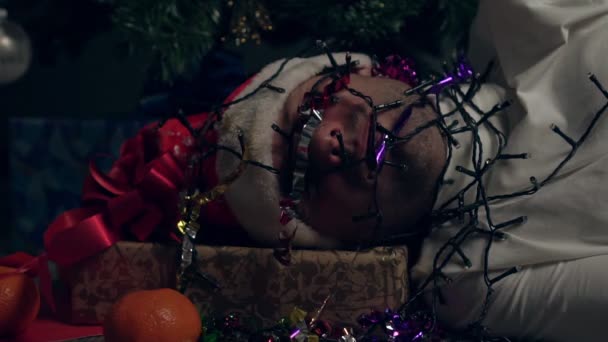 Un hombre con un sombrero de Papá Noel duerme en casa bajo un árbol de Navidad. Un hombre con una guirnalda en la cara — Vídeos de Stock