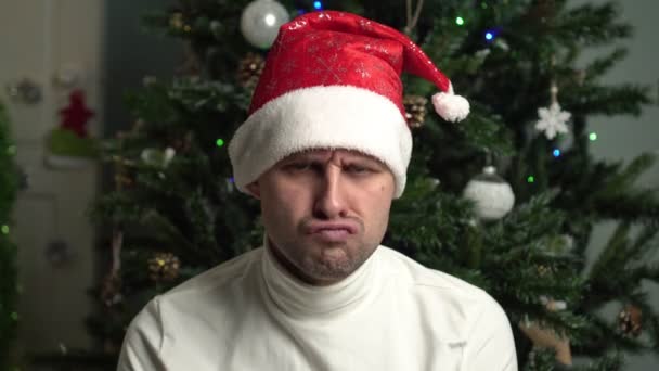 Θυμωμένος κακός άνθρωπος στο καπέλο του Αϊ Βασίλη στο φόντο του χριστουγεννιάτικου δέντρου. — Αρχείο Βίντεο