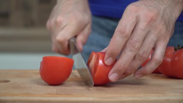 Ένας άντρας κόβει μια φρέσκια κόκκινη ντομάτα με μαχαίρι. Ένας άνθρωπος ετοιμάζει λαχανικά, σωστή διατροφή. Έννοια χορτοφαγίας — Αρχείο Βίντεο
