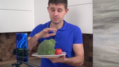 Erkek şef video blogcusu mutfakta yemek pişiriyor. Bir yemek blogcusu akıllı telefondan sebzelerle ilgili bir video çekiyor