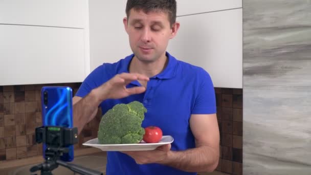 Мужчина шеф-повар видео блоггер готовится на кухне. Блогер о еде снимает видео об овощах на смартфоне — стоковое видео