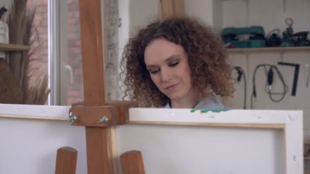 Un jeune artiste aux cheveux bouclés dessine un tableau dans un atelier d'art. Femme artiste heureuse au travail — Video