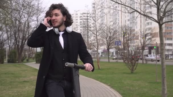 Modern üzletember beszél egy okostelefonon a szabadban. Egy férfi elektromos robogóval a parkban.. — Stock videók