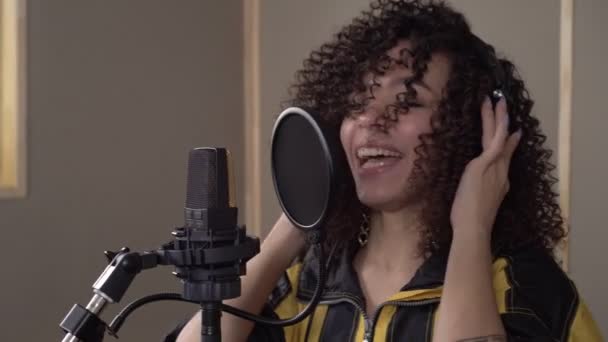 Счастливая прекрасная черная певица в наушниках поет в микрофон в студии звукозаписи. — стоковое видео