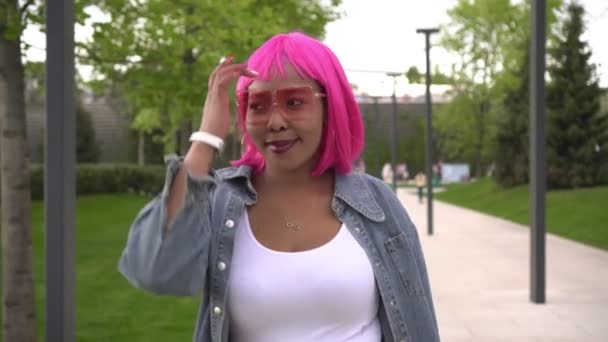 Pembe saçlı şık siyahi bir kadın güneşli bir günde parkta yürüyor. — Stok video