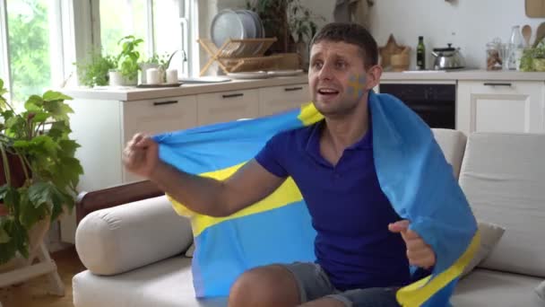 Appassionato di sport maschile a casa con la bandiera della Svezia. tifoso di calcio svedese guarda la TV e si rallegra per la vittoria — Video Stock