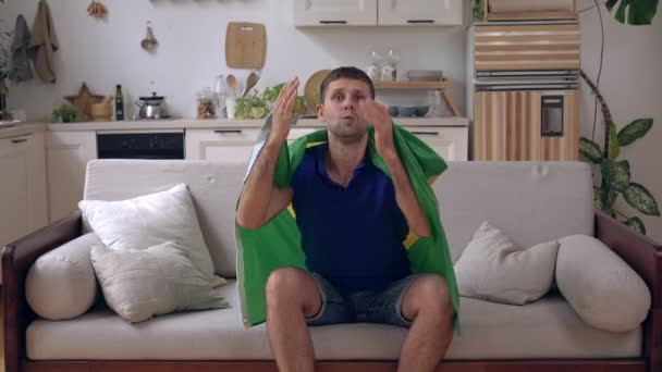 Appassionato di sport maschile a casa con bandiera brasiliana. Il tifoso brasiliano guarda la TV e gioisce della vittoria. — Video Stock