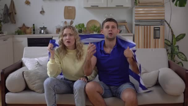 ギリシャの旗を持つ家庭でのスポーツやサッカーファンのギリシャの家族のカップル。ギリシャのファンはテレビを見ている. — ストック動画