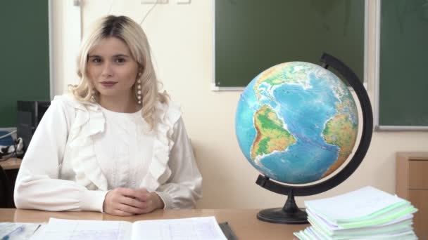 Portret van een jonge schoolleraar aan tafel in de klas met een wereldbol — Stockvideo