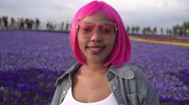 Genç, mutlu, siyah, pembe saçlı, mor çiçeklere gülümseyen bir Afrikalı kadın.