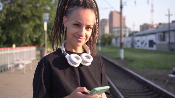 Mujer atractiva joven con teléfono inteligente en la estación de tren. Mujer moderna adolescente con rastas en el fondo del ferrocarril — Vídeos de Stock