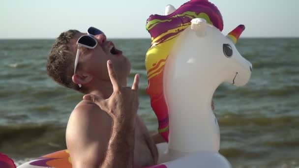 Смешной веселый мужчина с надувным плавательным кольцом в форме единорога на морском пляже — стоковое видео