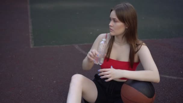 Młoda zmęczona kobieta nastolatka na boisku do koszykówki odpoczywa i pije wodę z butelki — Wideo stockowe