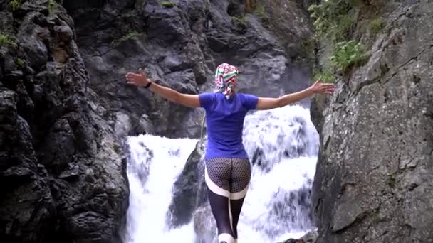 Una mujer está de pie sobre el fondo de una cascada. Mujer excursionista en una caminata cerca de la cascada — Vídeos de Stock