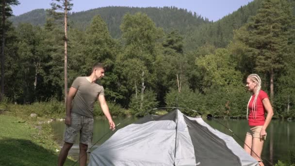 Egy férfi és egy nő túráznak, felállítanak egy sátrat. A fiatal turisták egy sátor a természetben, közel a tó az erdőben — Stock videók