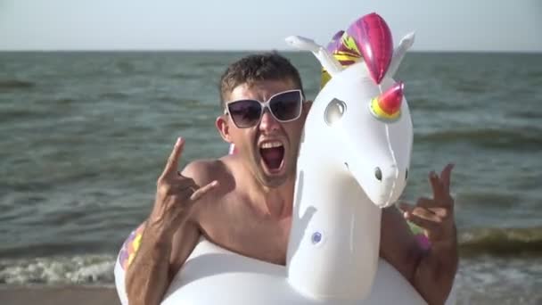 Amusant homme joyeux amusant à la mer. Un homme avec un anneau de natation en forme de licorne sur la plage — Video