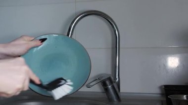 Bir ev kadınının elleri mavi bir tabak tutar ve lavabonun üstündeki musluktan akan suyun altında bir fırçayla yıkar. Yakın çekim, ev rutini, günlük hayat.