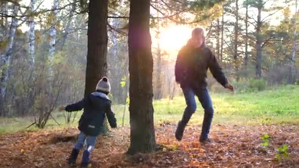 Un adolescente juega con un niño, corre por el parque alrededor de un árbol y lo alcanza. Los rayos del sol penetran en el bosque de pinos. Risa y alegría de toda la familia. — Vídeo de stock