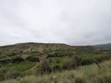 A breathtaking view of a mountain valley covered in tough grass and rocks. The light suggests changing weather, contrasting the bright sky with the deep green slopes.