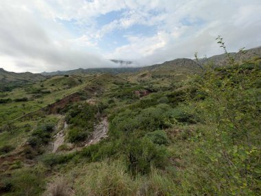 Layers of hills recede to the horizon under an overcast sky. The foreground is filled with scrub, stones, and a splash of autumnal color, showcasing the wild beauty of the region.
