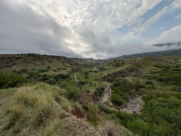 A breathtaking view of a mountain valley covered in tough grass and rocks. The light suggests changing weather, contrasting the bright sky with the deep green slopes.
