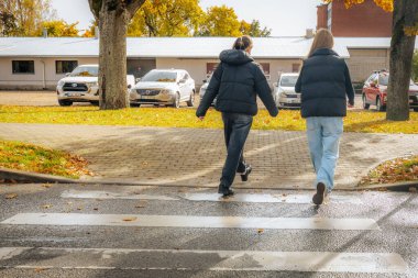 Siyah kıyafetli iki kişi, düşen sonbahar yaprakları, park edilmiş arabalar ve kentsel ve doğal elementlerin karışımı ile çevrili ıslak bir yaya geçidini geçiyor.. 