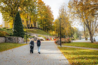 Sırt çantalı iki kişi, sonbahar yaprakları, ortanca çiçekleri ve arka planda sarı bir bina ile dolu bir parkta ağaçların oluşturduğu patika boyunca yürür..
