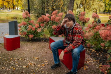 Rengarenk bir sonbahar parkında, ekose ceketli genç bir adam kırmızı bir bankta oturuyor. Etrafı pembe ortancalarla çevrili ve boynunda bir kamera var.. 