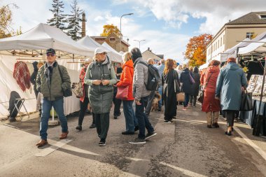 İnsanlar, beyaz çadırlar ve renkli ağaçlarla çevrili, hareketli bir sonbahar pazarında yürürler, ceket giyerler ve bulutlu bir gökyüzünün altında mevsimlik eşyalar için alışveriş yaparlar.. 