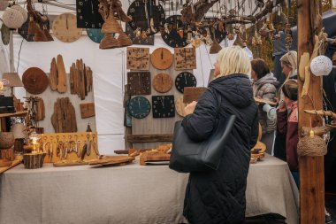 Bir grup insan el yapımı ahşap saatler, anahtarlıklar ve eşyalar satan bir el işi tezgahını araştırıyor. Sıcak ışıklandırma ve kırsal tasarımlar rahat ve sanatsal bir atmosfer yaratır..