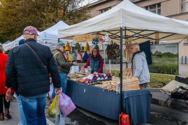 Pazarda beyaz bir gölgeliğin altında çeşitli el yapımı ahşap mutfak malzemeleri sergileniyor. Bir müşteri malları incelerken, bir satıcı içeriden yardım eder..