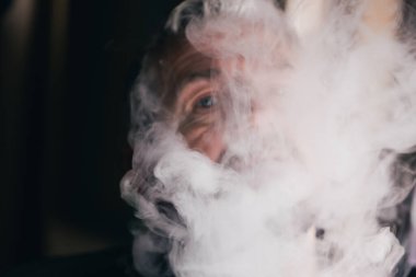 Kısmen örtülü bir adam yoğun duman ya da buharın içinden bakar, sadece bir gözü açıktır, gizemli ve kasvetli bir etki yaratır..